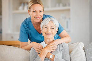A caregiver wearing blue scrubs crouching over to an elderly woman on a sofa