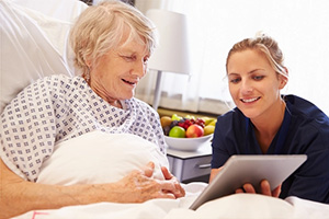 A caregiver wearing blue scrubs showing a computer to an elderly woman in bed