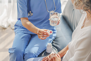 A nurse wearing blue scrubs providing medication to a resident sitting on a sofa